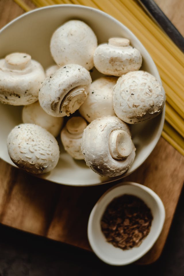 Fresh white button mushrooms in a white bowl beside dry spaghetti and a small dish of spices on a wooden board.